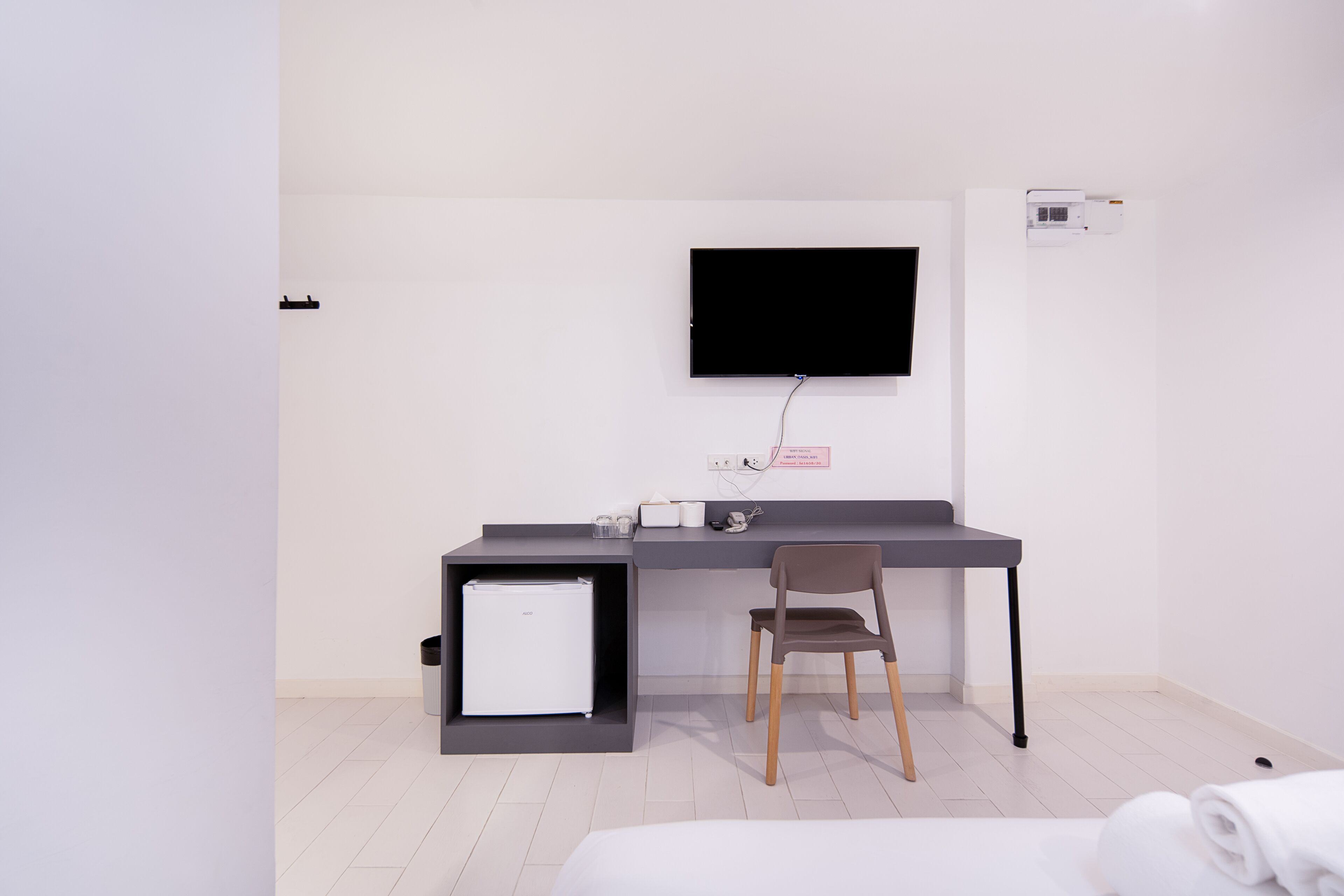 A modern hotel room with a bed, a desk with a chair, a flat-screen TV mounted on the wall, a small white refrigerator, and a wall-mounted air conditioning unit.