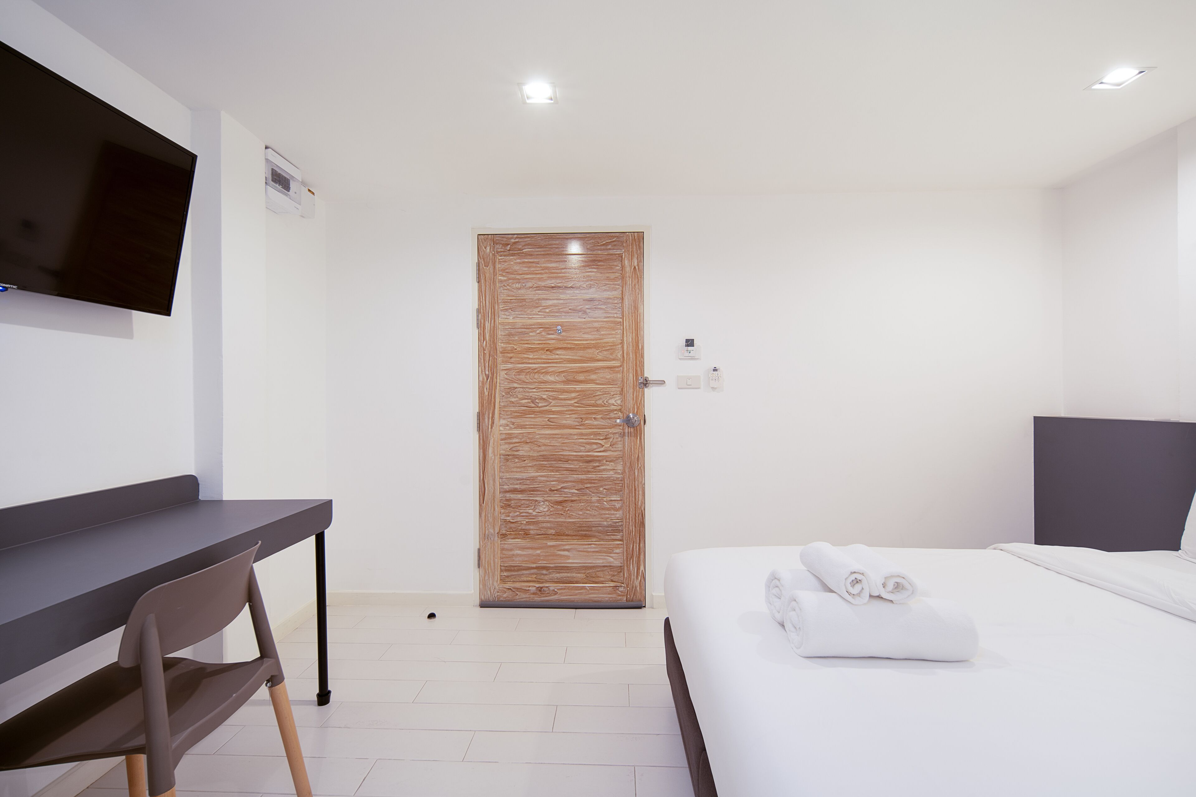 Habitación de hotel moderna con puerta de madera, televisor de pantalla plana en la pared, escritorio y una cama bien tendida con toallas enrolladas.