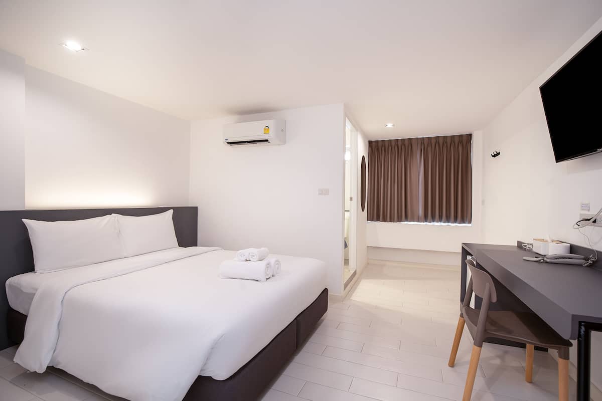 A hotel room with a bed, a desk, a flat-screen TV, an air conditioner, and a window with curtains.