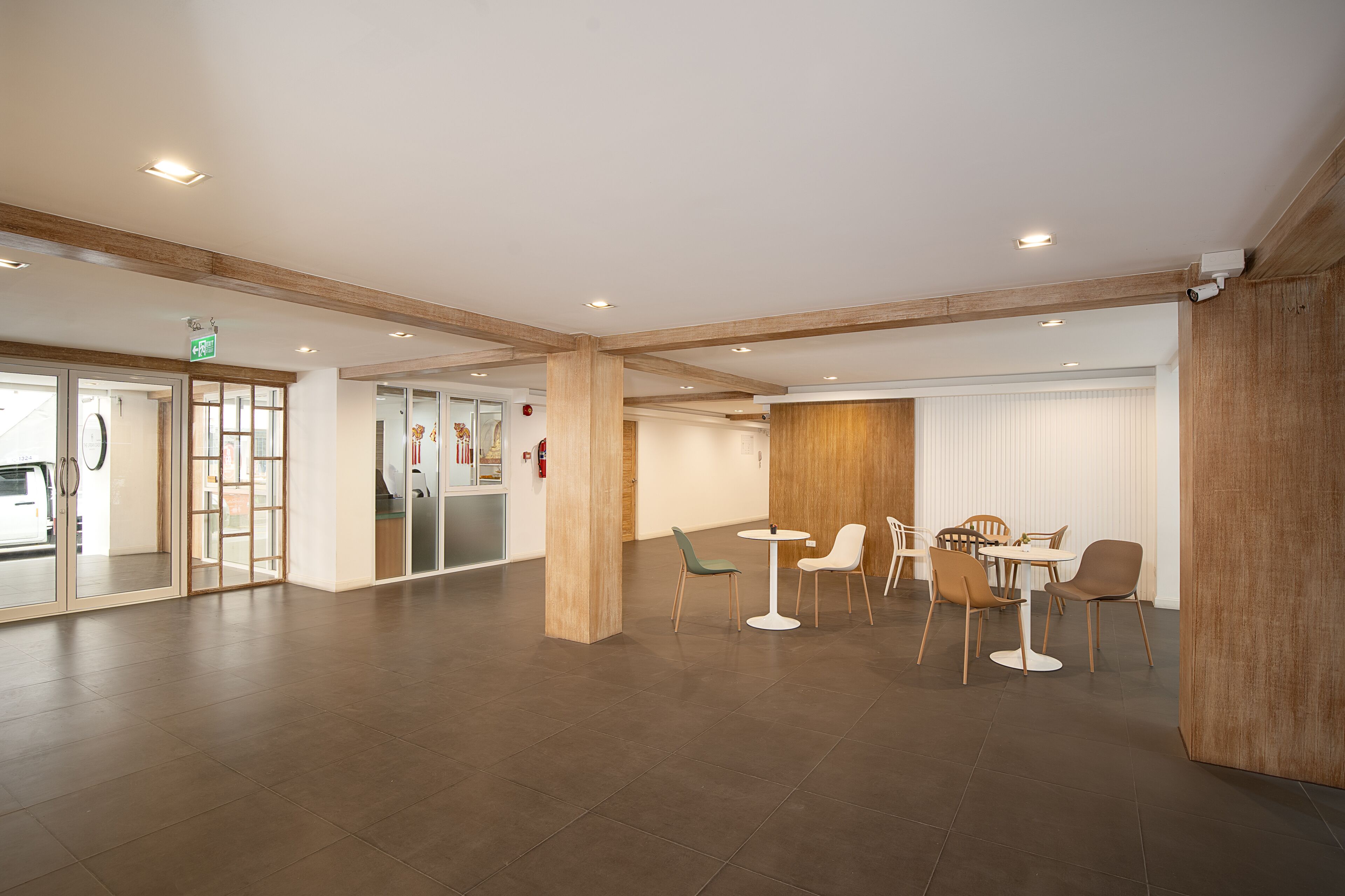 A spacious lobby with a modern design, featuring a central seating area with chairs and a table, wooden beams on the ceiling, and a glass door entrance.