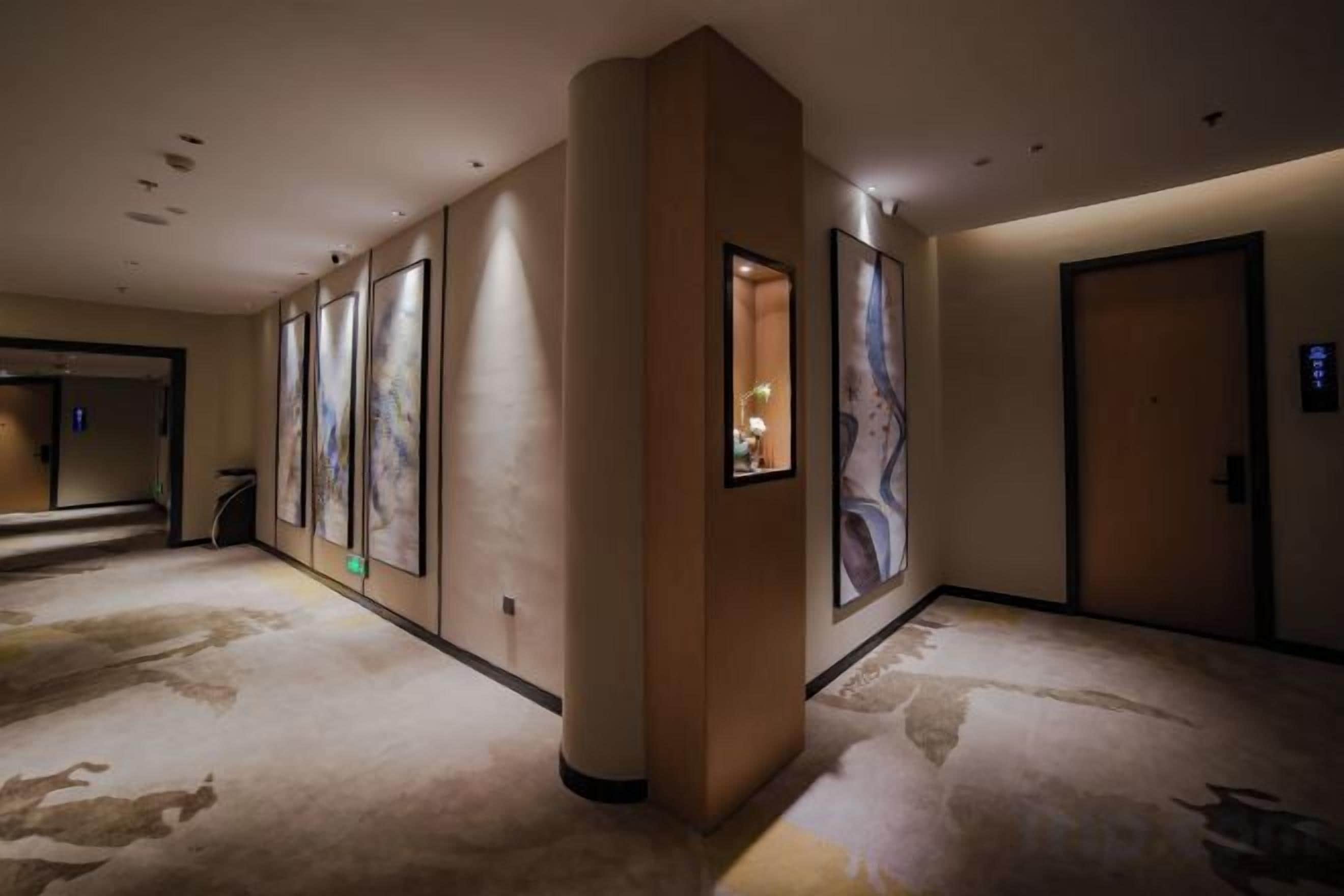 Corredor de hotel com quadros nas paredes, piso coberto por carpete e uma porta à direita.