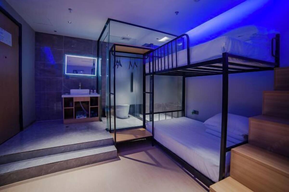 Habitación de hotel compacta con literas, baño con paredes de vidrio y una pequeña zona de escritorio.