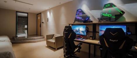 A modern hotel room with a gaming setup including two monitors, a gaming chair, and a desk.