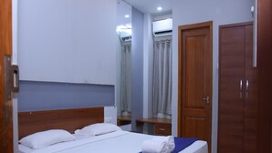 Ein ordentlich bezogenes Bett mit weißen Textilien und blauem Akzent, ein Holzschrank und ein Spiegel, der ein Badezimmer mit Duschvorhang reflektiert.