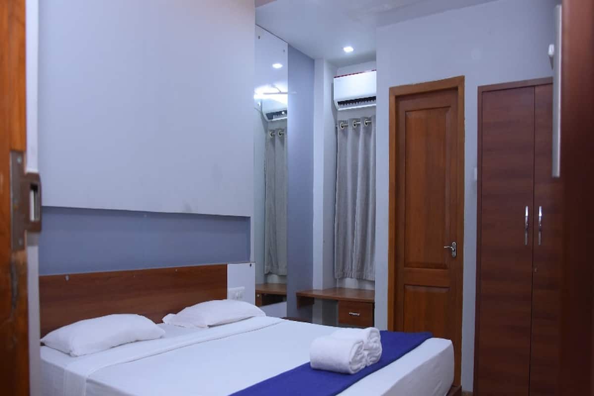 A neatly made bed with white linens and a blue accent, a wooden wardrobe, and a mirror reflecting a bathroom with a shower curtain.