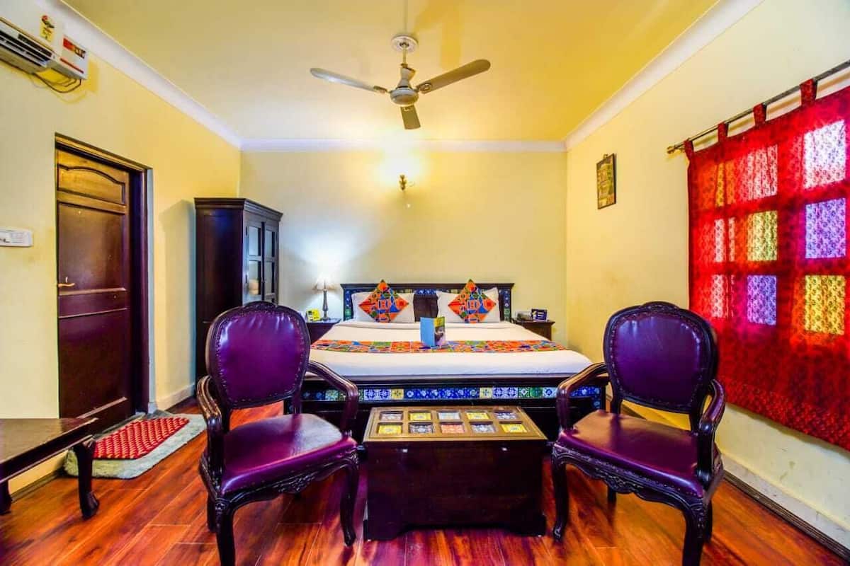 Ein Zimmer mit einem Bett, zwei violetten Stühlen, einem kleinen Tisch, einem Deckenventilator und einem roten, gemusterten Vorhang.
