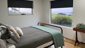 A bedroom with a bed, two windows with black blinds, a bedside table with a lamp, and a view of the sea. - Mount View Accommodation Marrawah (Marrawah)
