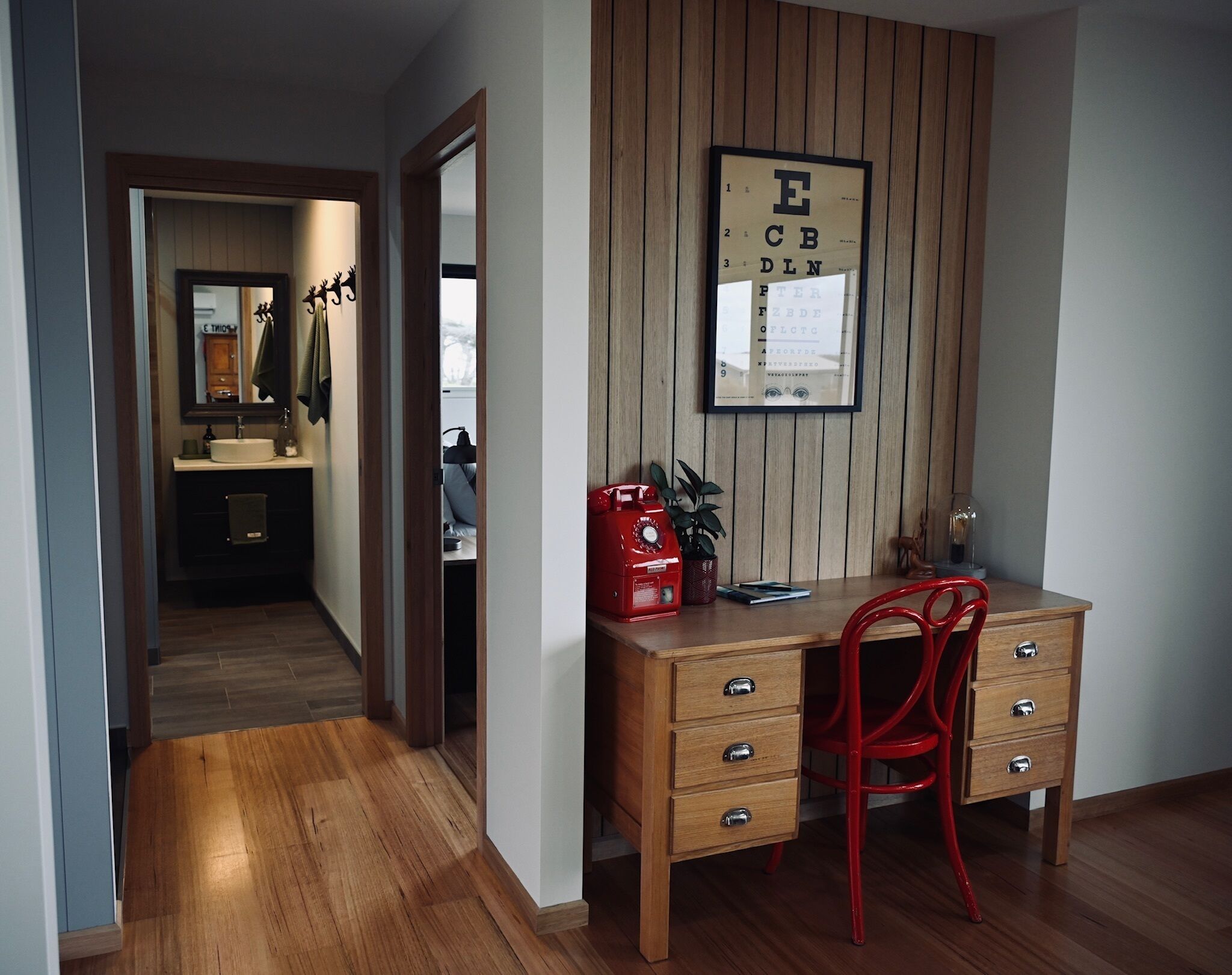 A study room with a wooden desk, a red chair, a framed eye chart on the wall, and a mirror above the desk.