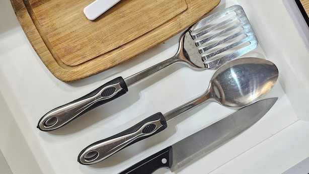 Un tiroir contenant une planche à découper en bois, un pinceau blanc et trois ustensiles de cuisine (une spatule, une pince et un couteau).