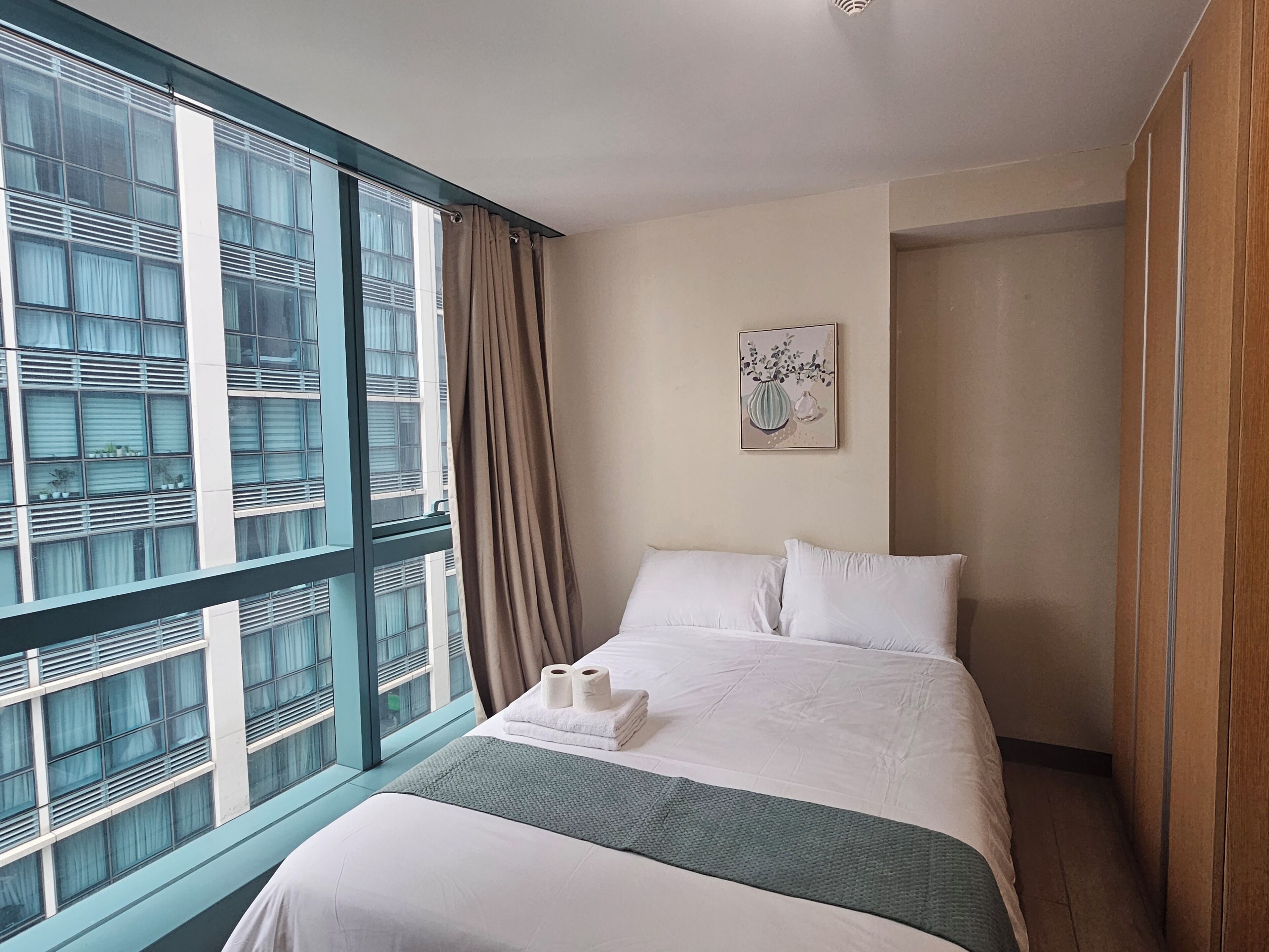 A hotel room with a large window, a bed with white linens, a green throw blanket, and a framed artwork on the wall.