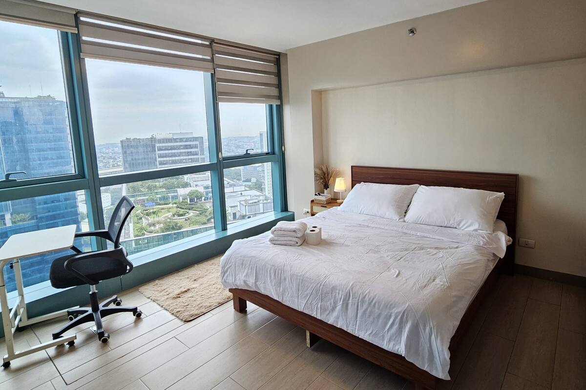 Quarto de hotel moderno com uma grande janela, cama de madeira, escrivaninha com cadeira e vista para a paisagem urbana.