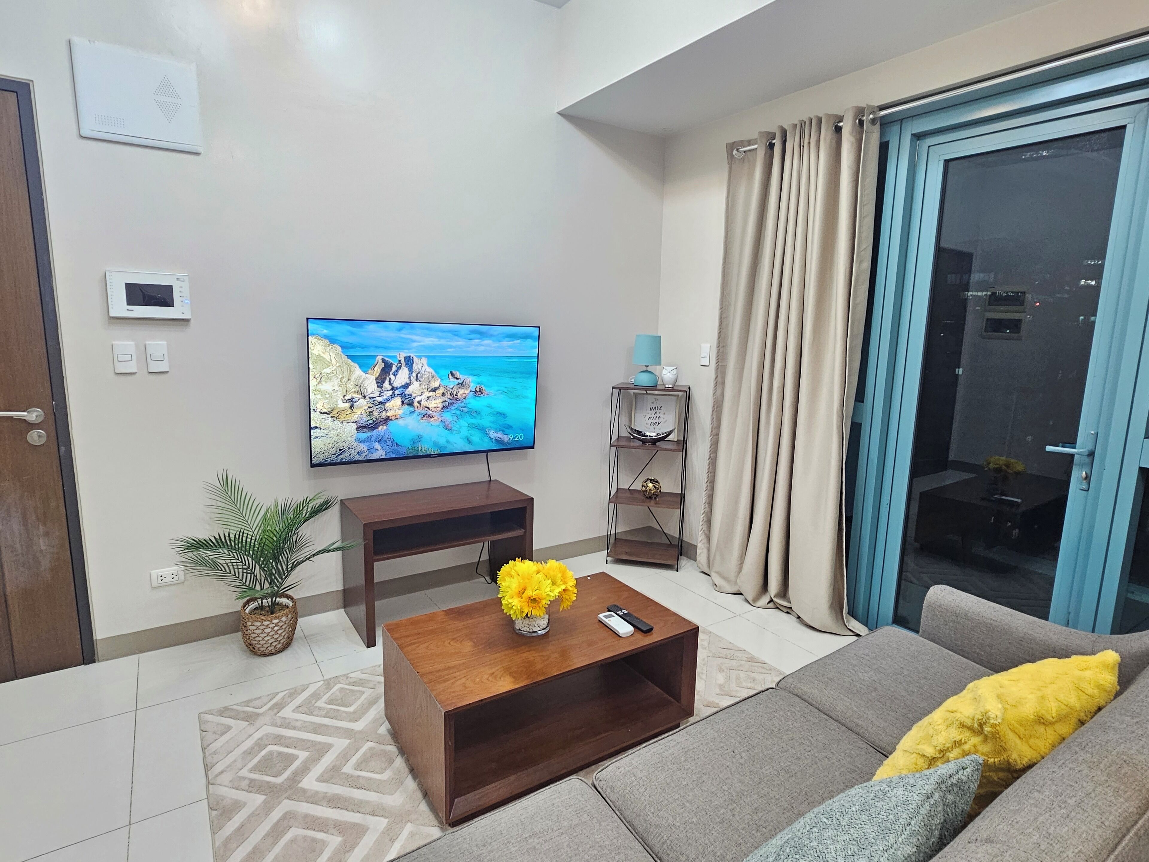 A living room with a flat-screen TV, a wooden coffee table with a vase of flowers, a grey sofa with yellow and grey cushions, a patterned rug, and a sliding glass door with curtains.