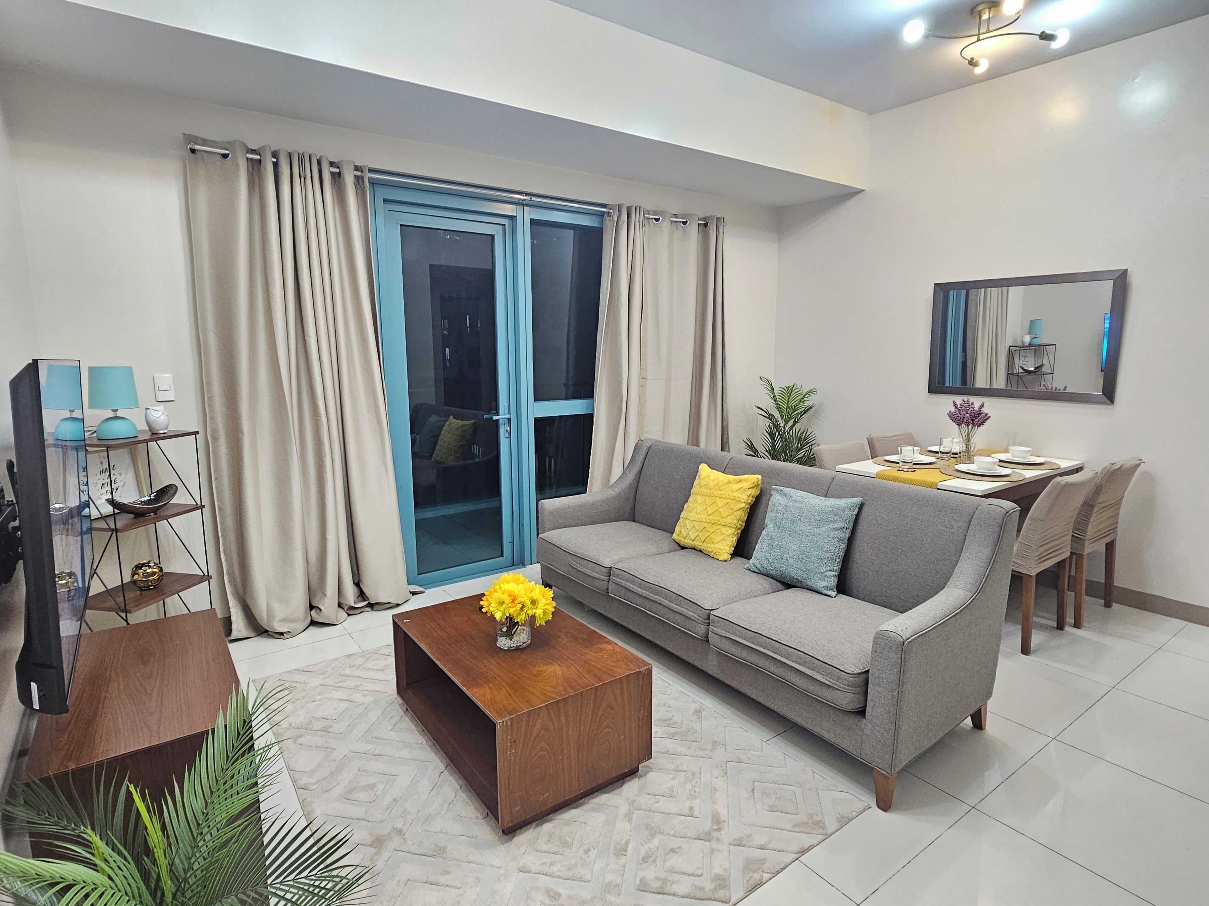 A modern living room with a grey sofa, yellow and blue cushions, a wooden coffee table with yellow flowers, a dining area with a table and chairs, and a sliding glass door with curtains.