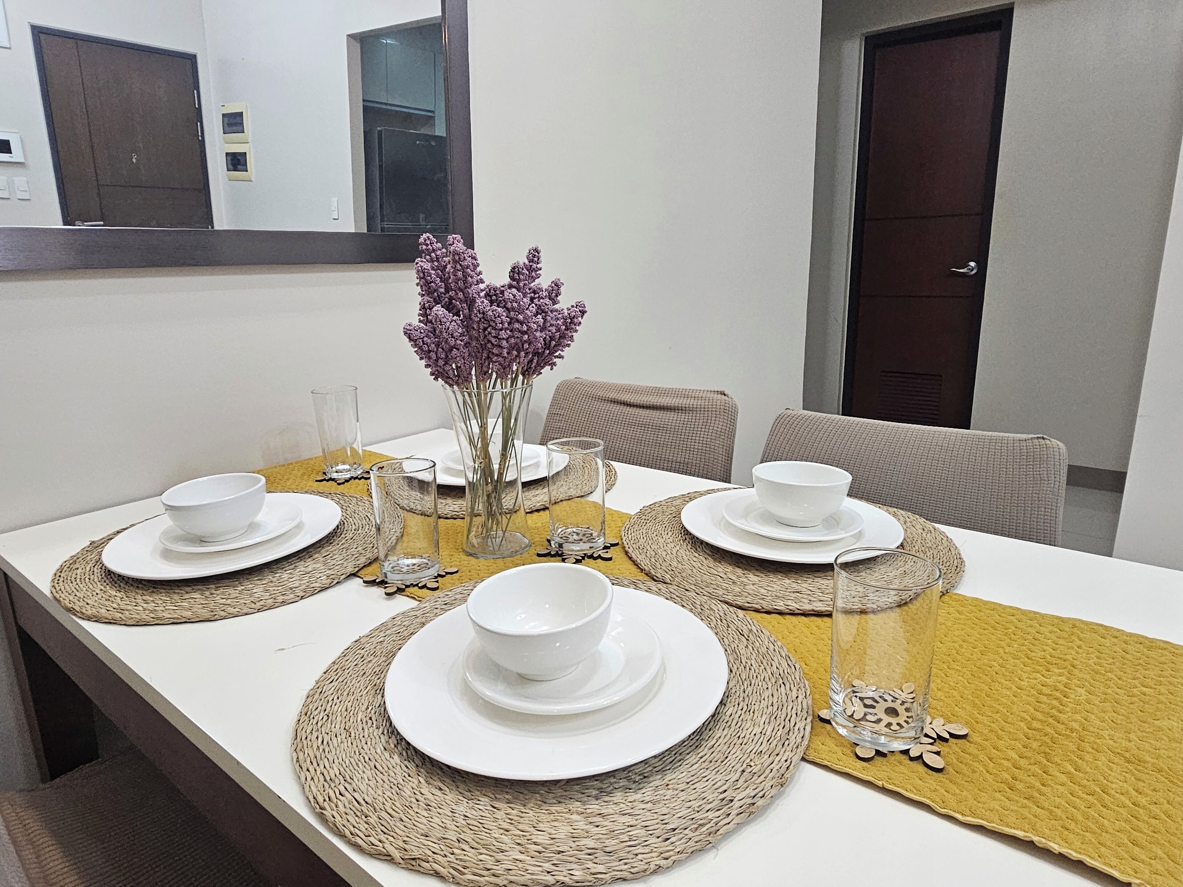 A dining table set with white plates, bowls, and glasses, surrounded by woven placemats and a vase with purple flowers.