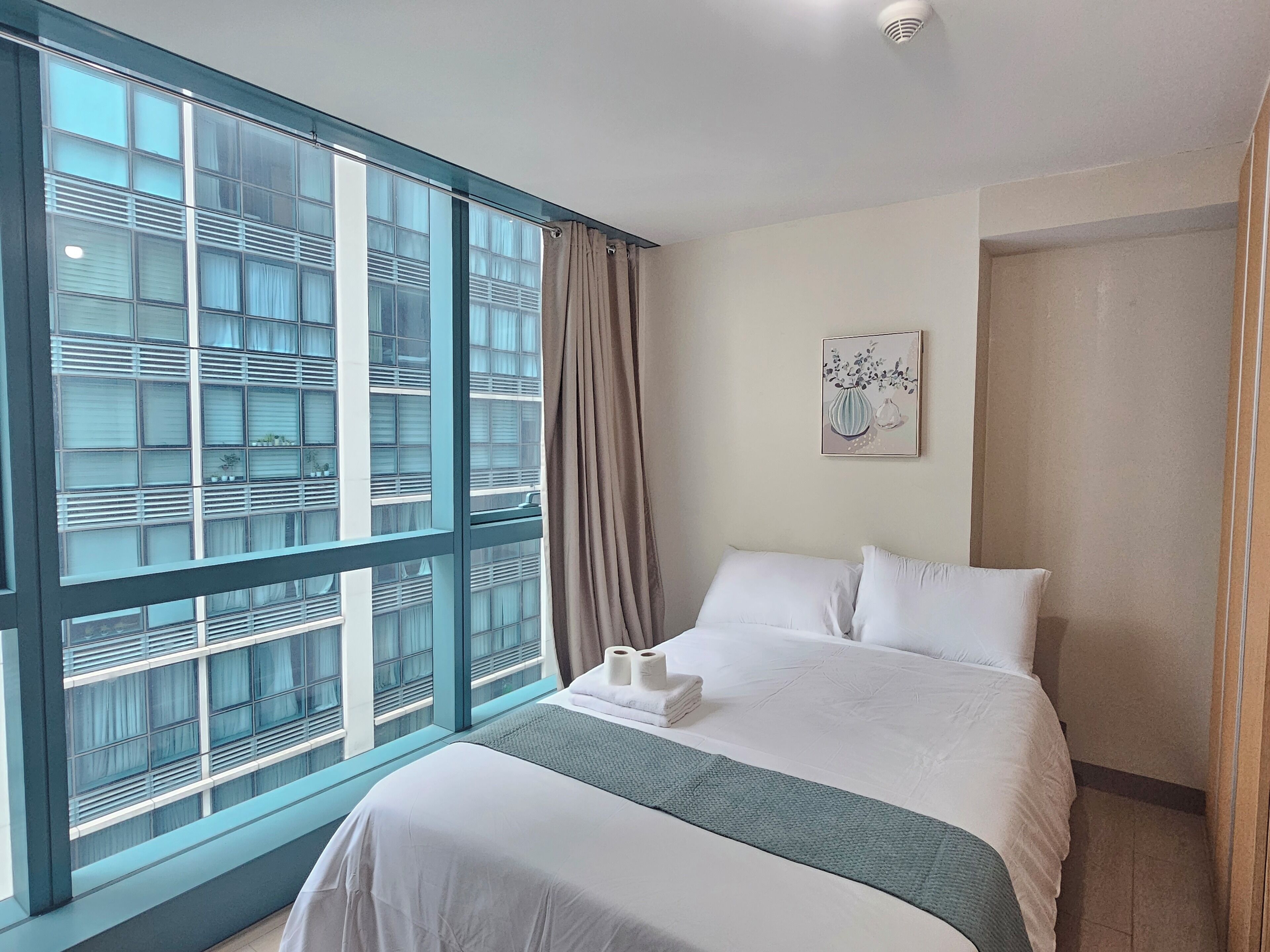 A hotel room with a large window, a bed with white linens, a painting on the wall, and a ceiling light.