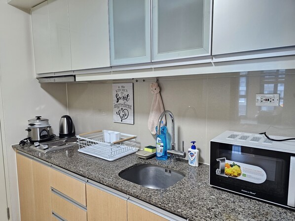 Uma cozinha com bancada de granito, pia de aço inoxidável, chaleira, escorredor de louça, micro-ondas e uma placa na parede.