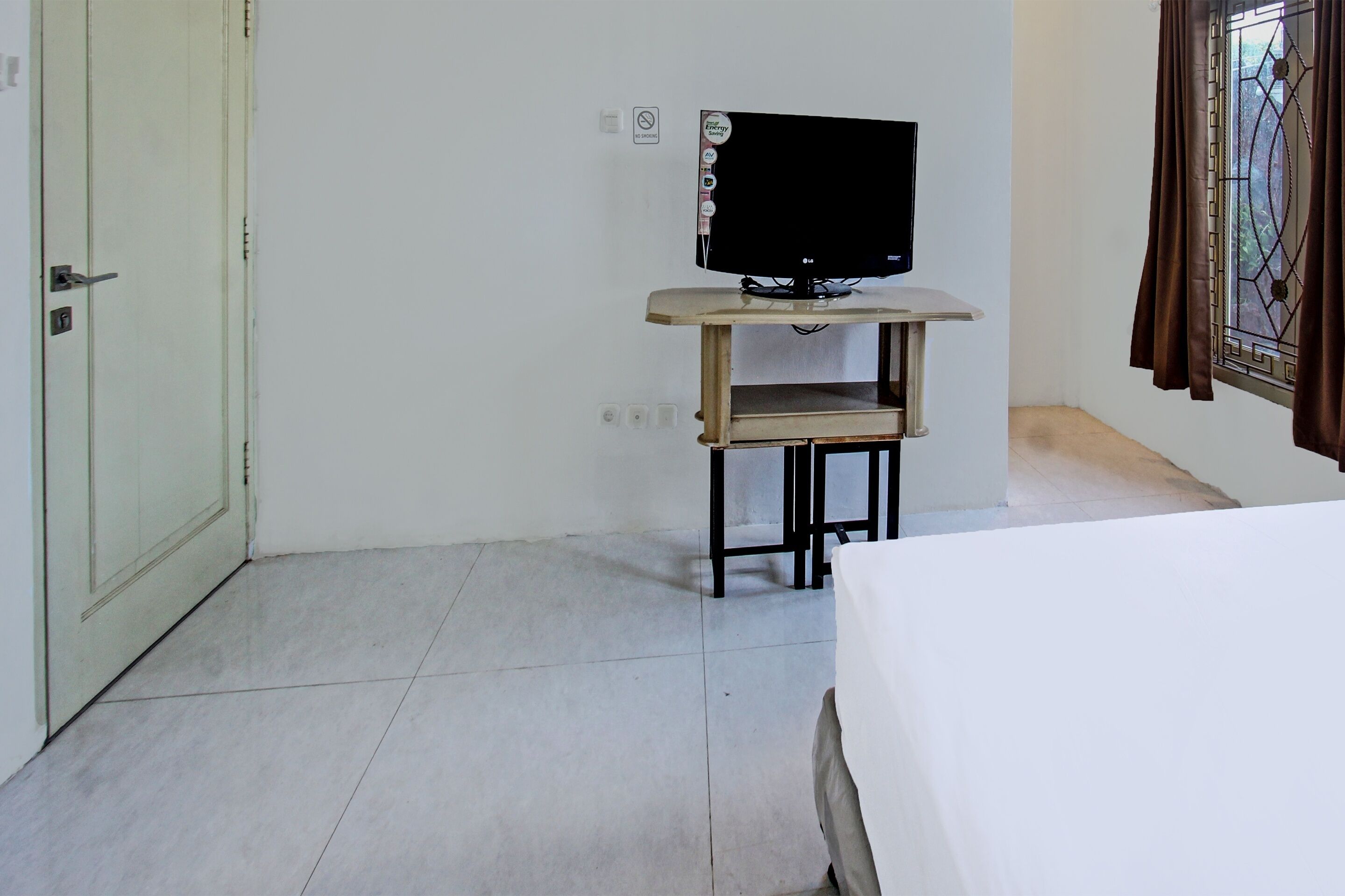 Una habitación con cama, televisor sobre un mueble de madera, puerta y ventana con cortinas.