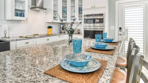 A kitchen with white cabinets, granite countertops, and a central island with blue bowls and plates. - Bougainvillea Beach Bungalow (St. Augustine)