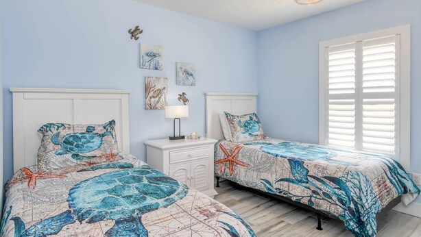 A bedroom with two beds, a ceiling fan, artwork on the walls, and a window with blinds. - Bougainvillea Beach Bungalow (St. Augustine)