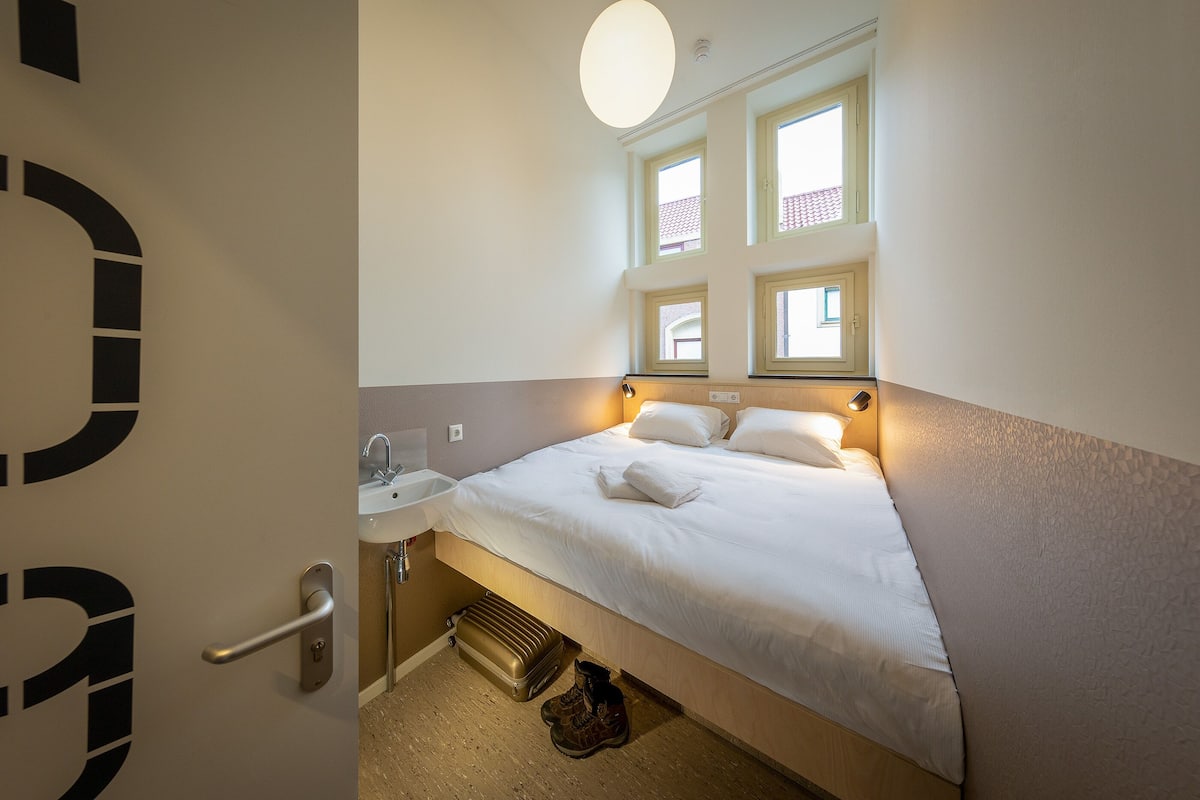 A hotel room with a bed, a window, a sink, and a door.