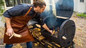 A person in a brown apron adding wood to a charcoal grill outdoors. - Stadsherberg Ald Weishoès (Venlo)