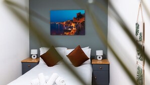 Ein ordentlich bezogenes Bett mit zwei braunen Kissen, ein Nachttisch mit Lampe und ein gerahmtes Bild mit einer Küstenlandschaft an der Wand.
