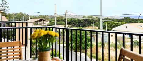 A wooden table set for dining with a vase of yellow flowers, plates, and a glass of water on a balcony with a view of the city and greenery.