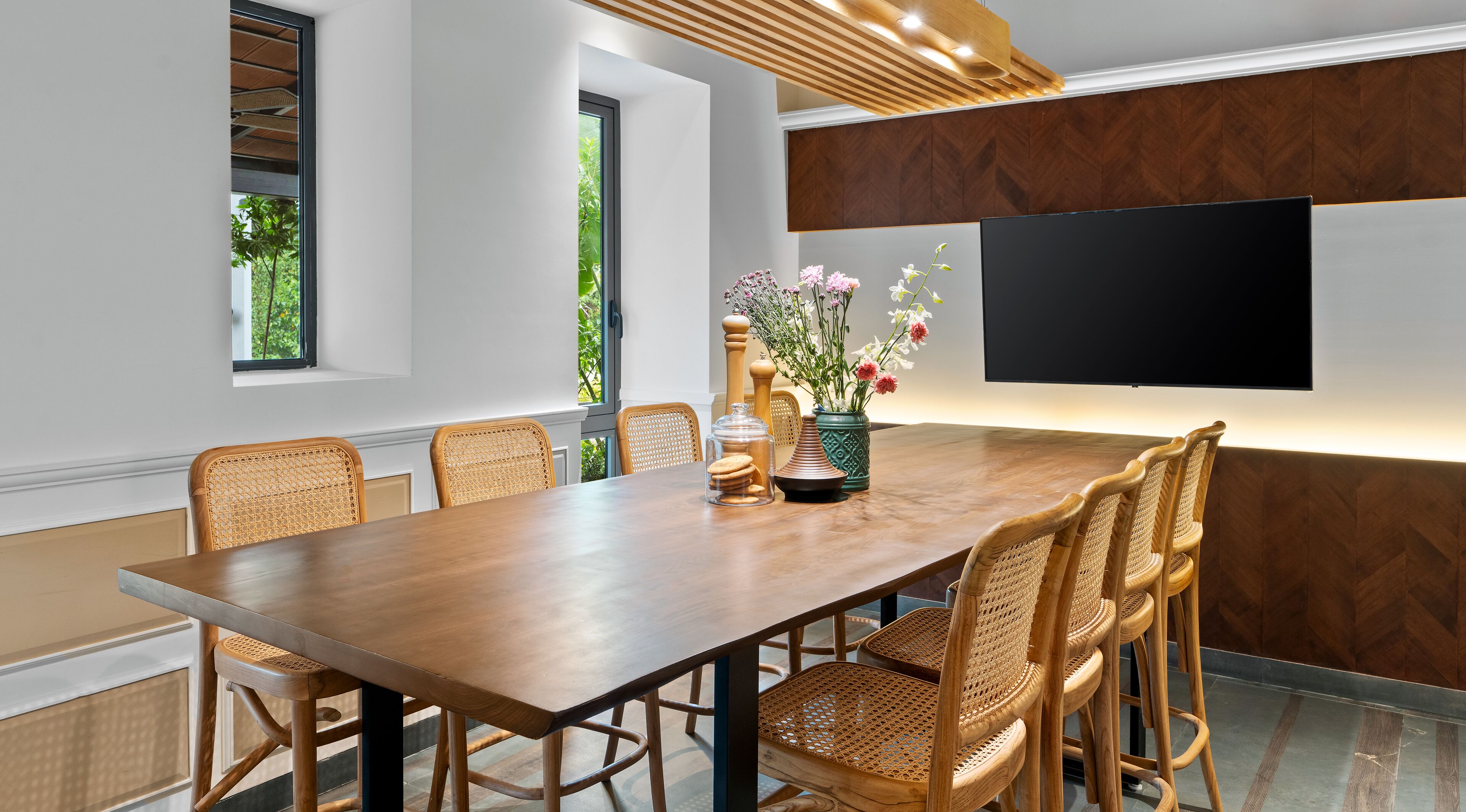 A modern dining area with a wooden table, wicker chairs, a vase with flowers, and a flat-screen TV mounted on the wall.