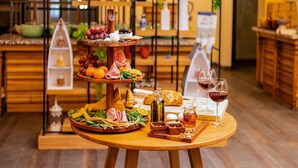 Una mesa de madera con un recipiente escalonado para servir, que contiene varios quesos, frutas y pan, junto con copas de vino y una botella de miel.