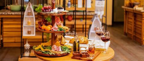 Una mesa de madera con un recipiente escalonado para servir, que contiene varios quesos, frutas y pan, junto con copas de vino y una botella de miel.