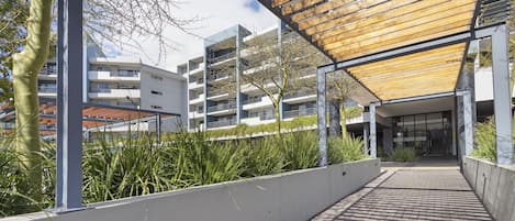 Un pasillo cubierto con techo de madera, una pared blanca con un jardín vertical y un edificio con varios balcones al fondo.