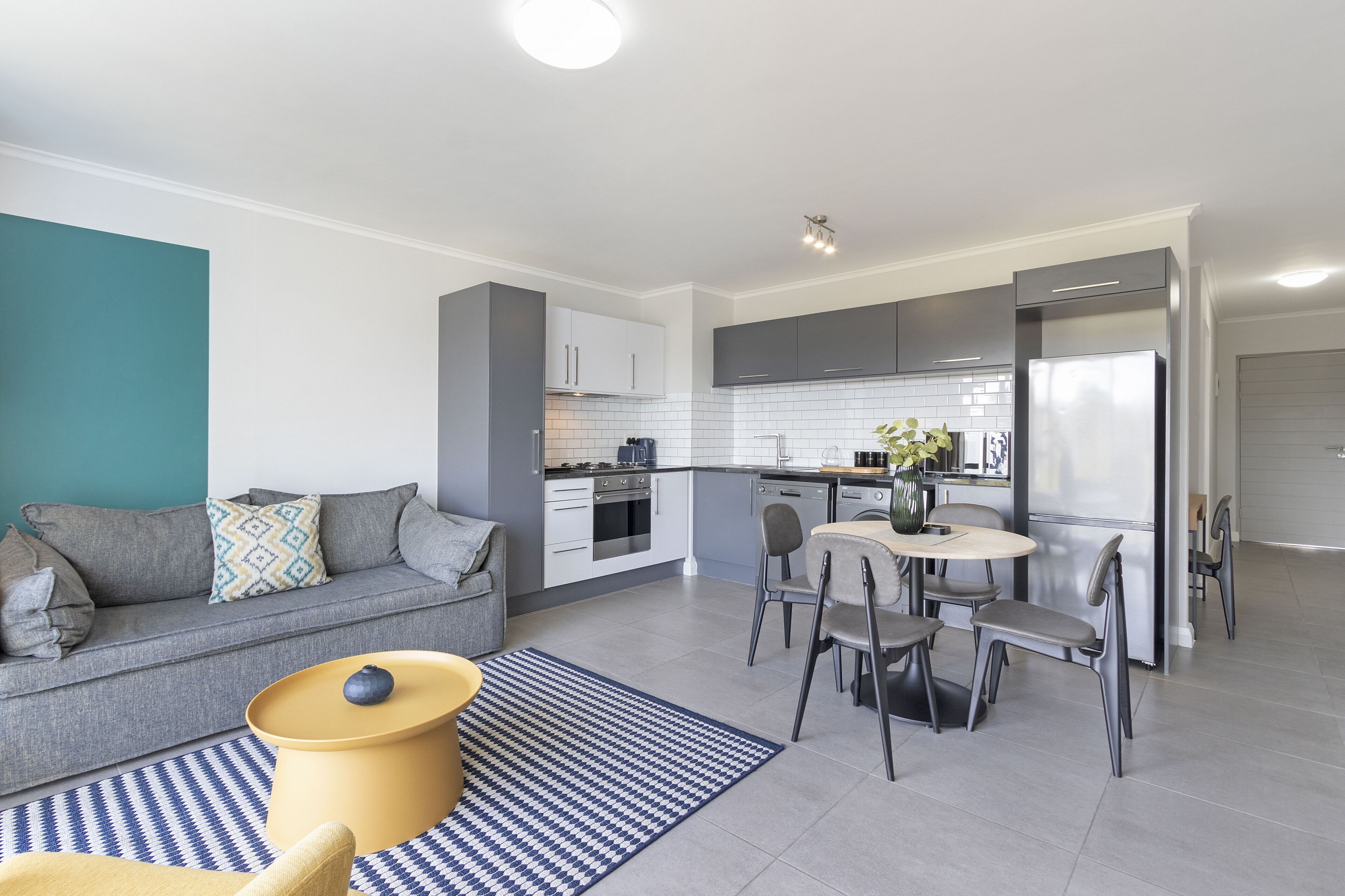 A modern kitchen with white cabinets, a dining area with a round table and chairs, and a grey sofa with patterned cushions.
