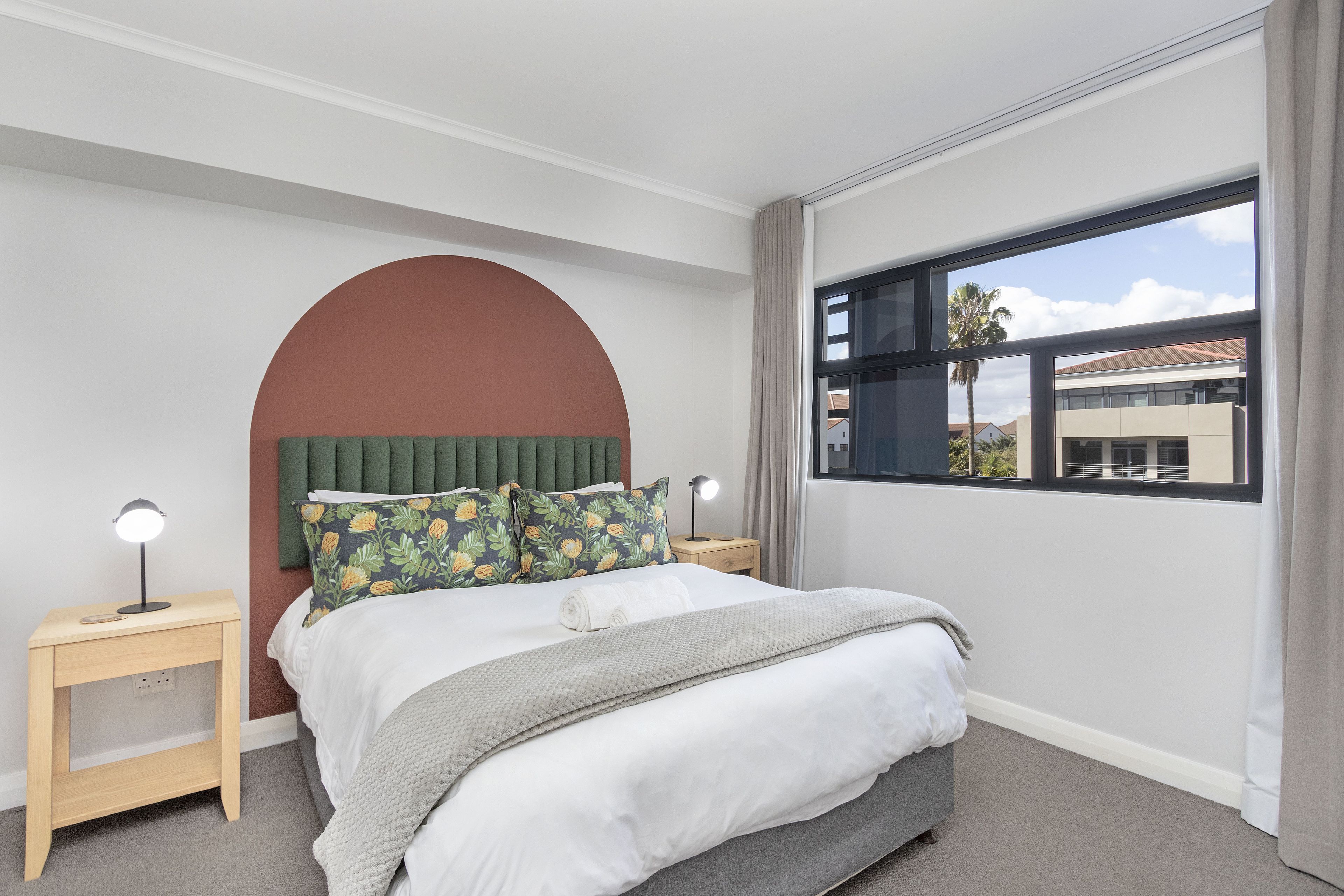 A bedroom with a bed, two nightstands, a large window, and a view of buildings outside.
