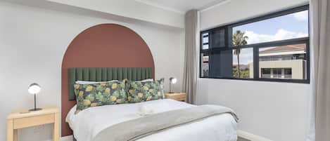A bedroom with a bed, two nightstands, a large window, and a view of buildings outside.