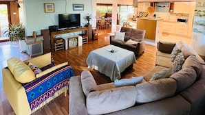 A spacious living area with a large sofa, a coffee table, and a television. There is a dining area with a table and chairs, and a kitchen visible in the background. - The Farm House
 (Possum Brush)