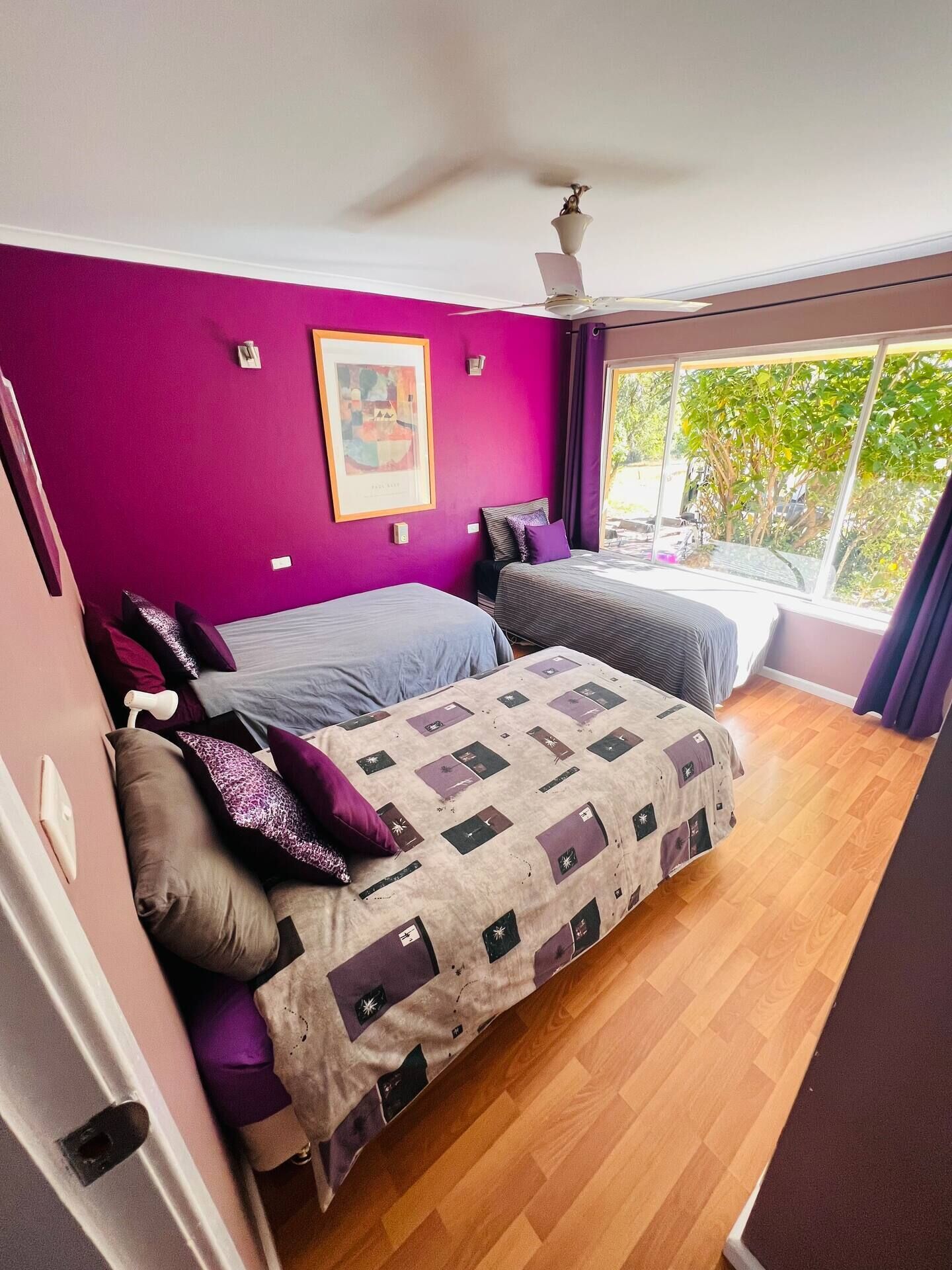 Ein Schlafzimmer mit zwei Betten, violetten Wänden, einem Deckenventilator, einem Fenster mit Aussicht und einem gerahmten Bild an der Wand.
