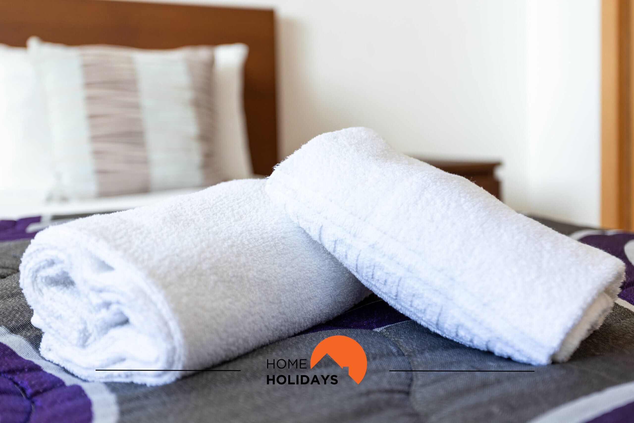 A neatly made bed with white towels, a wooden headboard, and a purple and white patterned bedspread.