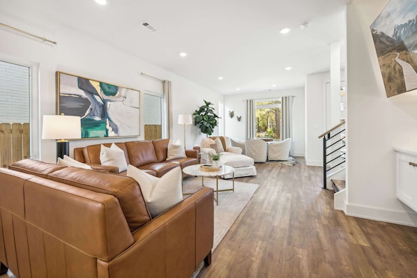 Un salon moderne avec un canapé en cuir marron, un fauteuil blanc et une table basse. Un tableau orne le mur, et un escalier mène à l’étage supérieur.