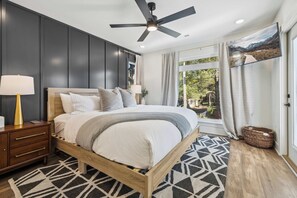Une chambre à coucher comprenant un lit, des tables de chevet, un ventilateur de plafond, une grande fenêtre avec des rideaux et une vue sur l’extérieur.