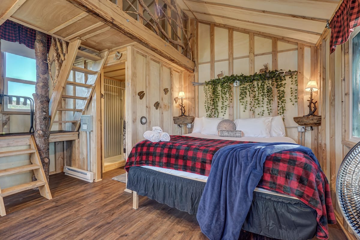 A wooden cabin bedroom with a bed, a ladder, a desk, and a window.