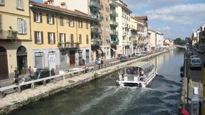 A canal with a boat, buildings along the water, and people walking on the sidewalk. - Navigli Suites (Milan)