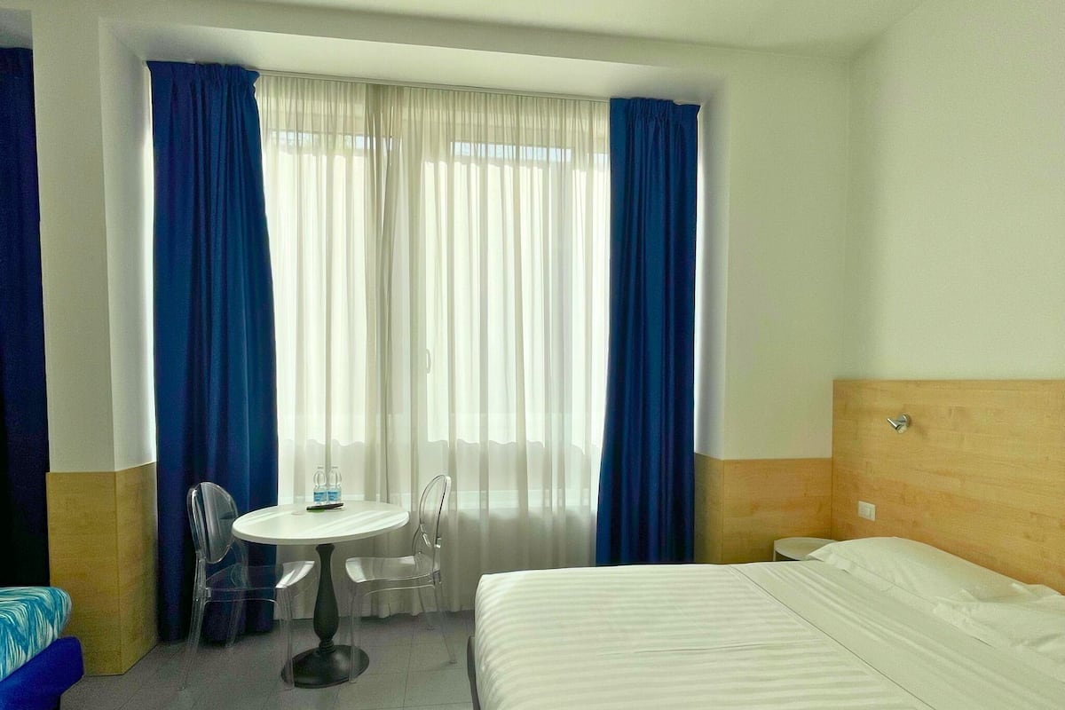 A hotel room with a bed, a round table with chairs, a small table with water bottles, and a window with blue curtains.