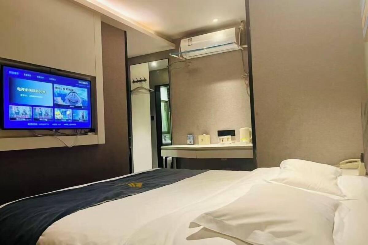 A hotel room with a bed, a TV mounted on the wall, a mirror, and a small table with a phone and a cup.