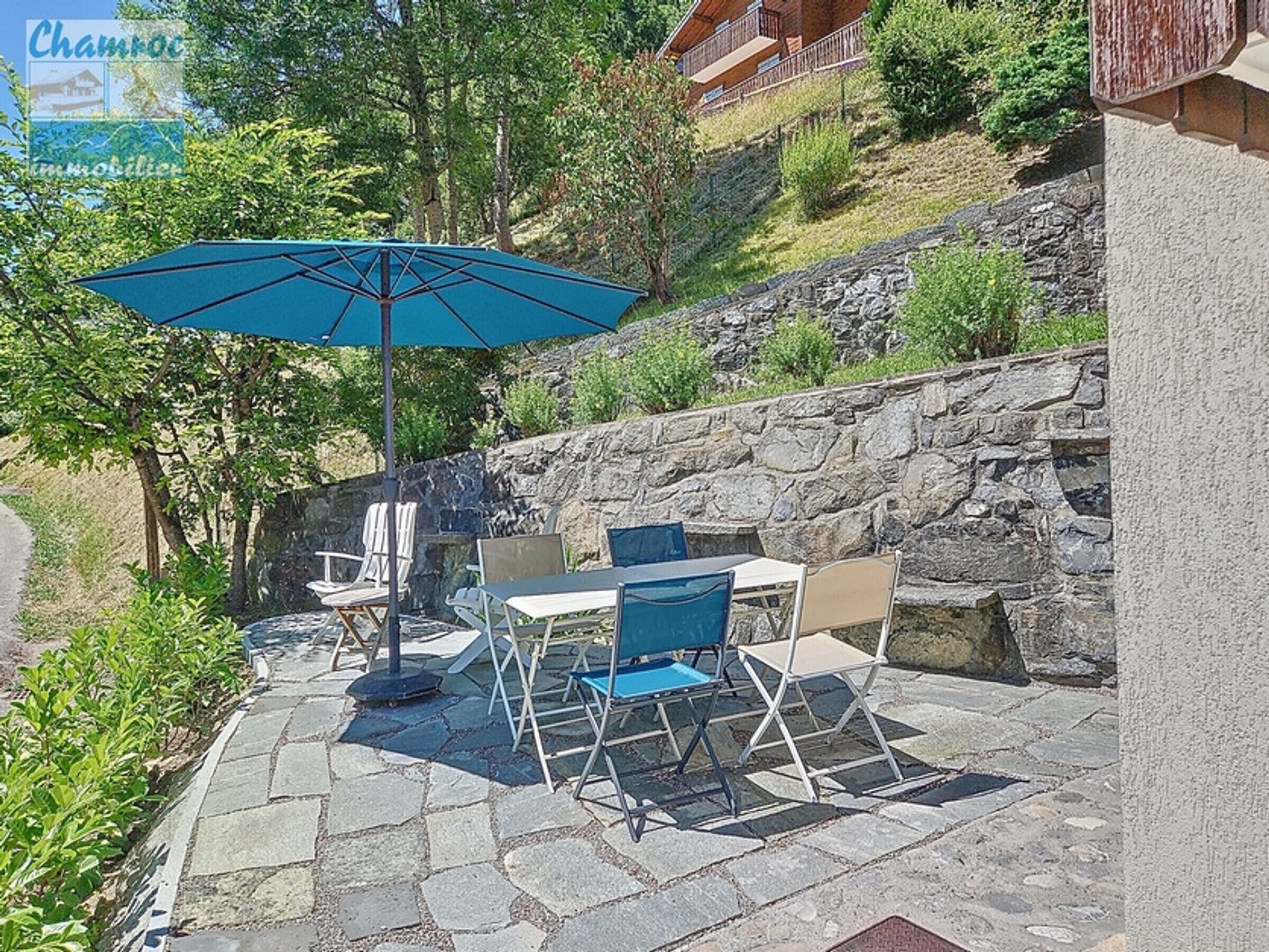 An outdoor patio with stone flooring, a table, chairs, and a blue umbrella.