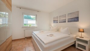 A modern bedroom with a large bed, a bedside table with a lamp, a window with blinds, and artwork on the wall. - Küstenliebe - Moderne und Gemütliche Wohnung mit Weitläufiger Terrasse (Koserow)
