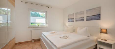 A modern bedroom with a large bed, a bedside table with a lamp, a window with blinds, and artwork on the wall.