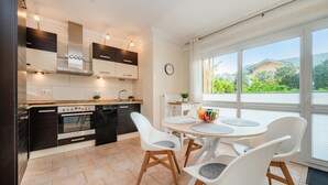 A modern kitchen with a built-in oven, a dining area with white chairs, and large windows offering a view of the outdoors. - Küstenliebe - Moderne und Gemütliche Wohnung mit Weitläufiger Terrasse (Koserow)