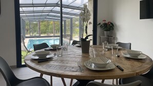 A dining area with a round table, chairs, and a view of a swimming pool through a glass enclosure. - Comfortable cottage with covered pool and spa in Couzon (Couzon)