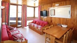 A cozy room with a wooden ceiling, a sofa with red cushions, a wooden dining table, and a television mounted on the wall.