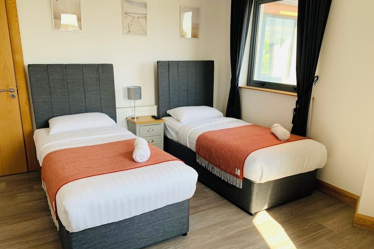 A hotel room with two beds, a wooden floor, a window with curtains, and a door.